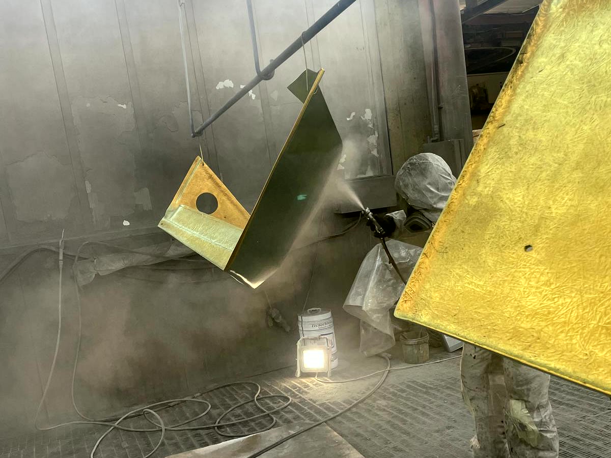 Operatore che vernicia a spruzzo un manufatto metallico appeso in cabina, accanto a un pannello già verniciato in giallo